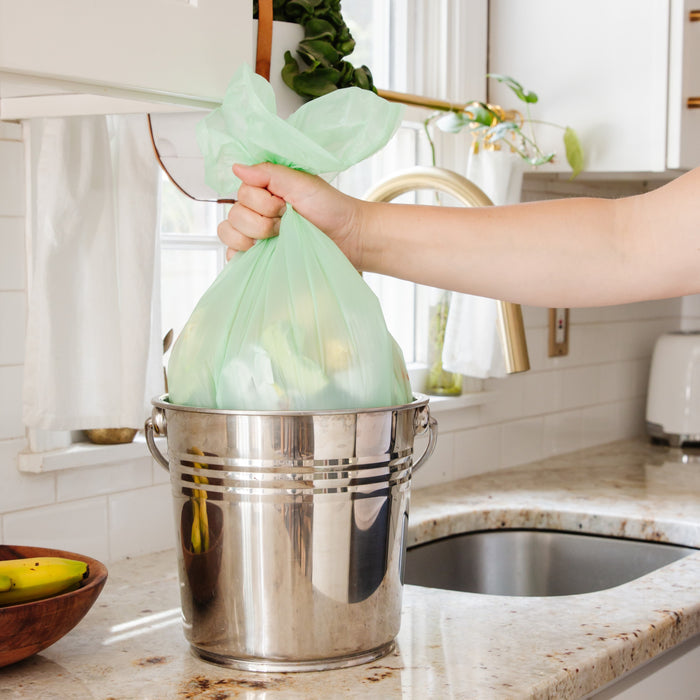 Someone places a tied Full Circle Compostable 4-Gallon Waste Bag into a stainless steel trash bin on the kitchen counter, which is next to a white sink, toaster, and a bowl of bananas.
