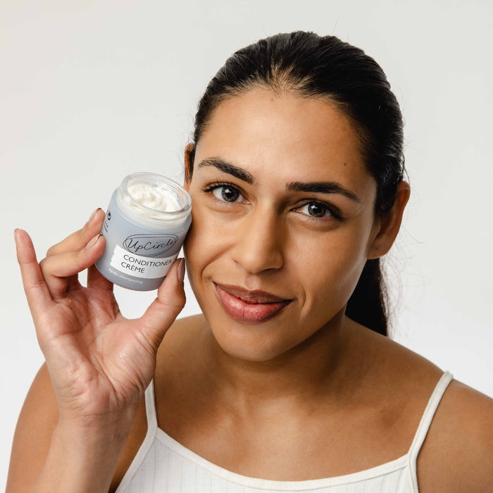 A person with long dark hair and a white tank top holds a jar of UpCircle Conditioner Crème enriched with rosemary oil, bamboo extract, and vitamin E, smiling slightly. The background is plain white.