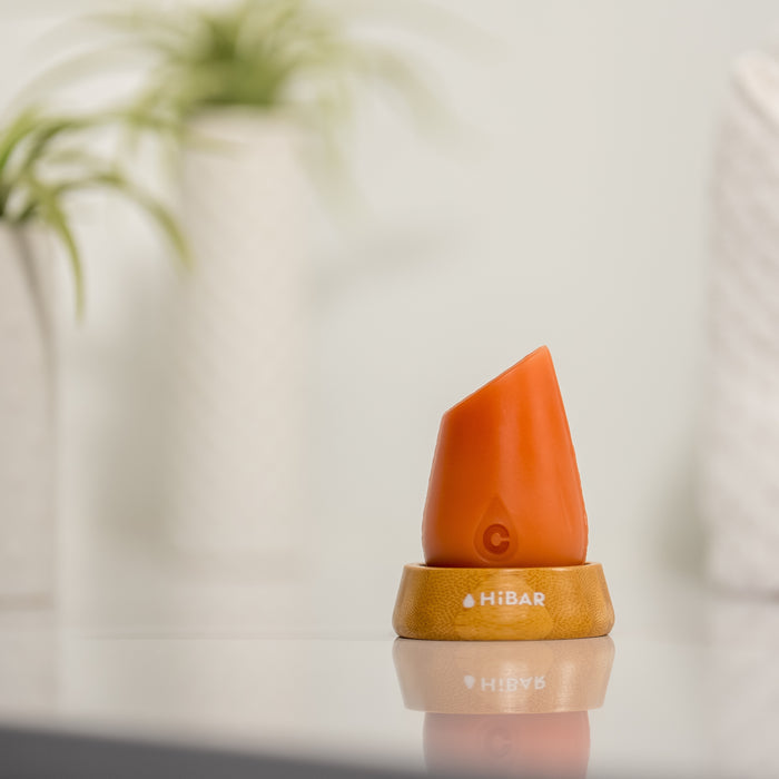 The HiBAR Curl Conditioner rests elegantly on a wooden stand on a reflective surface, flanked by two blurred green plants in white pots, creating a soft, natural ambiance perfect for curly hair care.