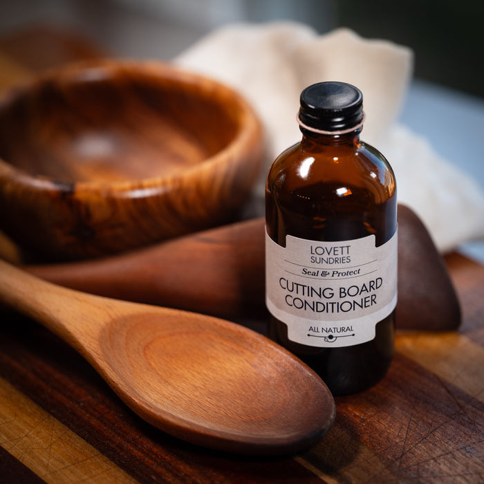 A brown glass bottle labeled Cutting Board Conditioner by Lovett Sundries sits on a wooden cutting board, showcasing coconut oil-based wood protection. Nearby are two wooden spoons and a bowl. The kitchen backdrop includes a white cloth.
