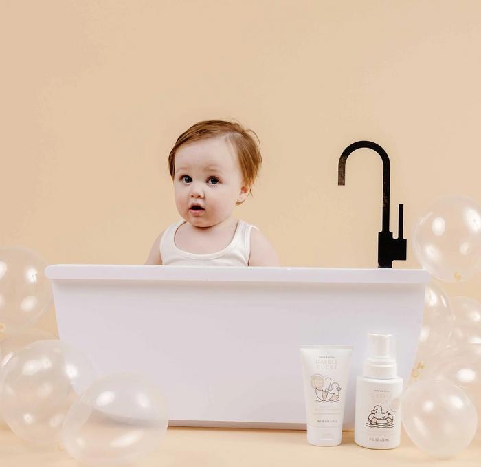 A baby with light brown hair sits in a white bathtub surrounded by translucent balloons, wearing a white tank top and looking surprised. Beside the tub are two bottles of Dabble & Dollops EWG-verified Dabble Ducky Infant Wash. The background is plain beige.