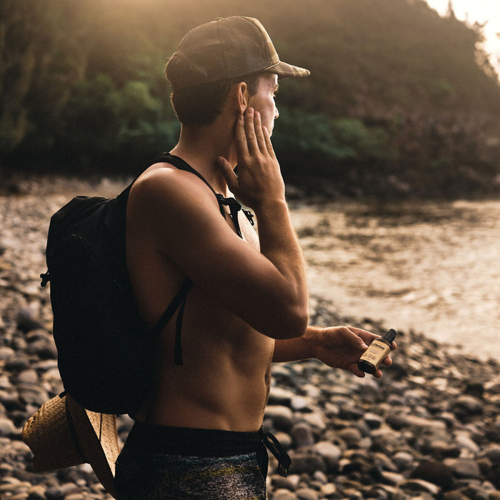 A man on a rocky beach applies Thrive Natural Cares Daily Sunscreen for Sensitive Skin - SPF 30 to his face, holding the bottle in one hand. Sunlight through trees provides UVA/UVB protection as he stands in a cap and backpack, facing the water.