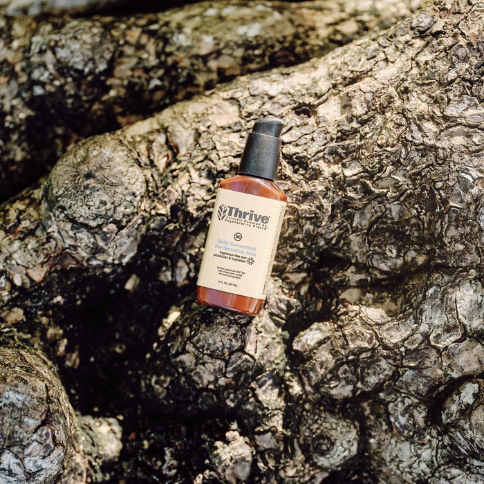A bottle of Thrive Natural Cares Daily Sunscreen for Sensitive Skin - SPF 30 sits on a textured, bark-covered surface. The amber-colored bottle with a black pump and beige label blends seamlessly with the earthy tones of the bark.