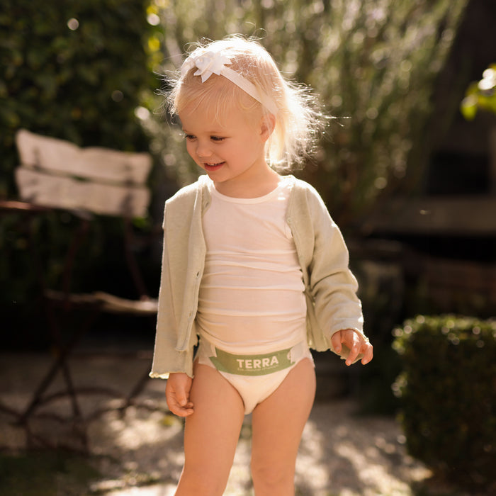 A blonde child wearing a white headband, a white shirt, and a light cardigan enjoys the outdoors, smiling in the sunlight amid greenery. The comfort from their Terra diapers plant-based materials is evident.