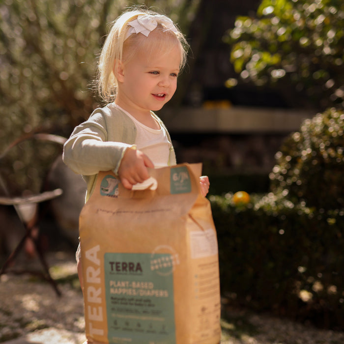 A young child outdoors smiles holding a large brown bag labeled Terra Diaper featuring bamboo fiber. The background is filled with greenery and soft sunlight.