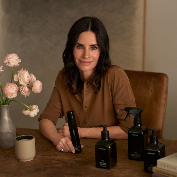 A dark-haired woman in a brown shirt sits at a table with pink flowers, a mug, and several black bottles of Homecourt Dish Soap made with plant-derived surfactants. She smiles softly and looks at the camera.