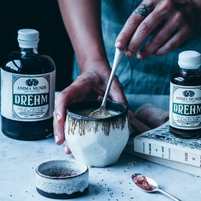 A person stirs a drink in a rustic ceramic mug beside jars of Anima Mundi DREAM Elixir, Sleep Aid + Third Eye Tonic, a small bowl of pink salt, and a book on a marble surface.
