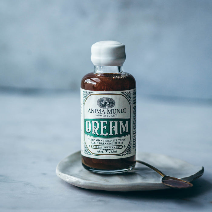 A small glass bottle labeled Anima Mundi DREAM Elixir, Sleep Aid + Third Eye Tonic, rests on a ceramic dish with a small spoon beside it, set against a neutral background.