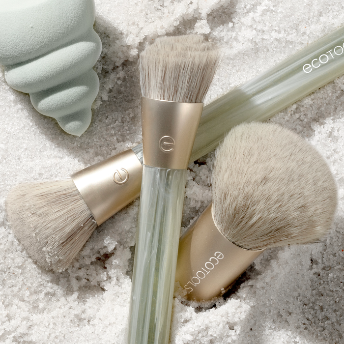 EcoTools Eco Sea Gems Face Set with light bristles and translucent handles lies on white sand. A mint green, cone-shaped object peeks from the top left. These cruelty-free brushes enhance your beauty routine with an eco-friendly touch.