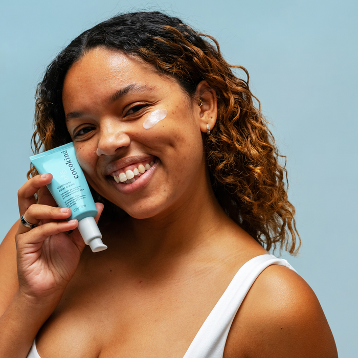A smiling woman with curly hair showcases a blue Cocokind Electrolyte Water Cream tube against a light blue backdrop. Dressed in a white tank top, she sports a dab of moisturizer on her cheek, exuding promises of deep hydration.