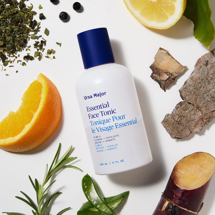 A white bottle of Ursa Major Essential Face Tonic is surrounded by natural ingredients like lemon, orange, rosemary, birch bark, sugarcane, and peppercorns on a white background. This exfoliating tonic helps promote clearer skin.