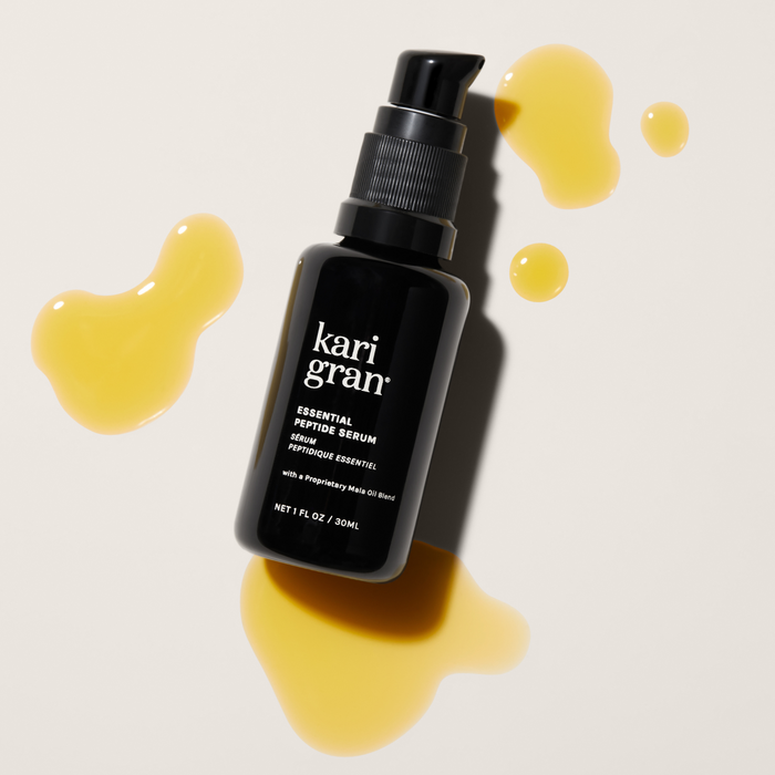 A black bottle of Kari Gran Essential Peptide Serum with a pump top sits on a light surface, surrounded by yellowish serum forming abstract shapes. It features Matrixyl 3000 and peptides, with 1 fl oz / 30 ml marked at the bottom.