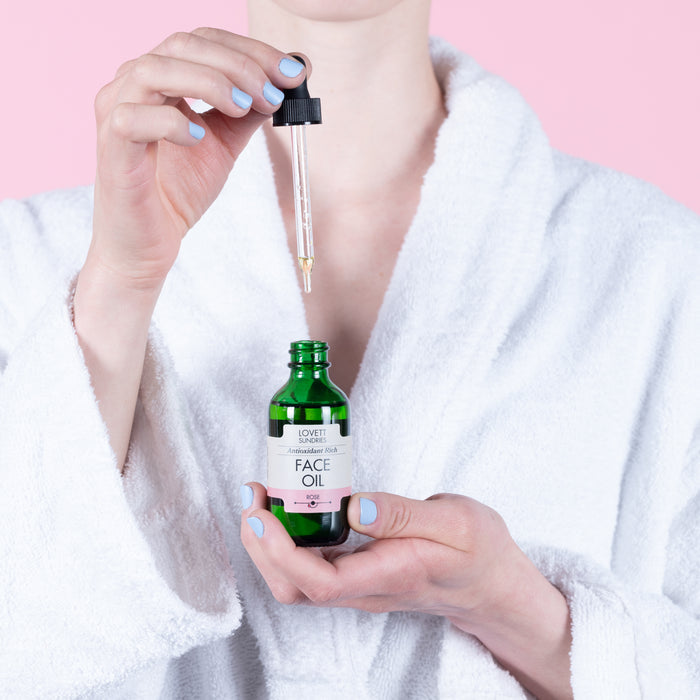 A person in a white robe holds a green bottle of Lovett Sundries Face Oil, complete with a dropper, against a pink backdrop. Their light blue-painted fingernails reflect the calming nature of this organic elixir designed to boost skin elasticity.
