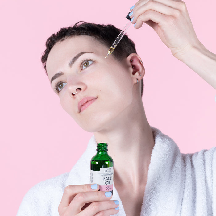 A person in a white robe applies a drop of Lovett Sundries Face Oil using a dropper. They hold the green bottle, renowned for enhancing skin elasticity with organic oils, against a vibrant pink background.