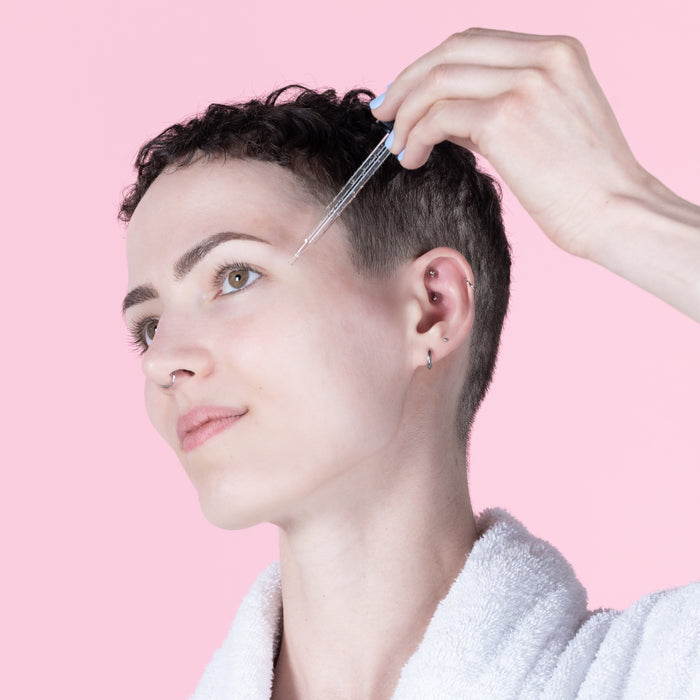 A person with short hair uses a dropper to apply Lovett Sundries Face Oil, enhancing skin elasticity. Theyre in a white robe with ear piercings against a soft pink background.