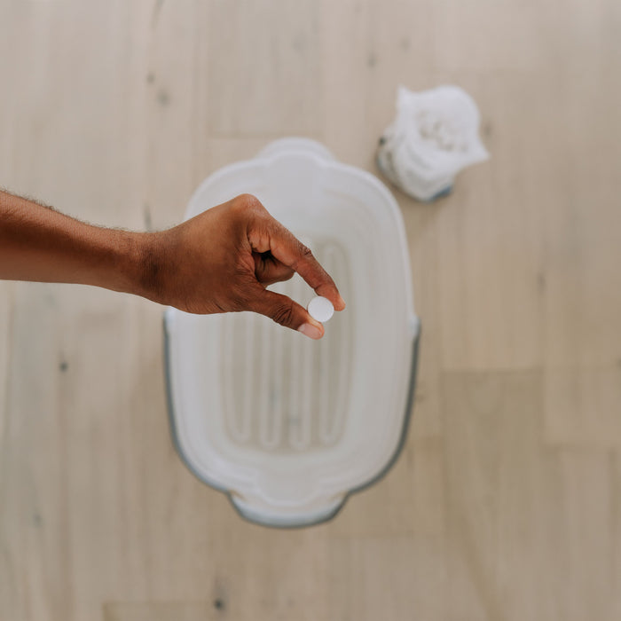 A hand holds a The Unscented Company Floor Cleaning Tab—fragrance and dye free—over an empty white basin on a light wooden floor, with white cloths in the background.
