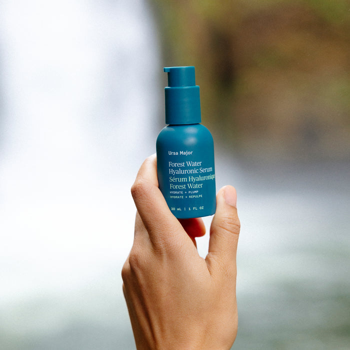 A hand holds a teal bottle of Ursa Majors Forest Water Hyaluronic Serum against a blurred waterfall, underlining its hydrating and purifying effect for dehydrated skin.