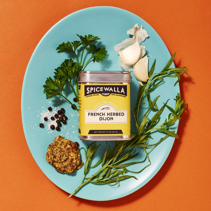 A blue plate holds a tin of Spicewalla French Herbed Dijon, surrounded by parsley, tarragon, garlic cloves, black peppercorns, sea salt, and whole grain mustard on an orange background.