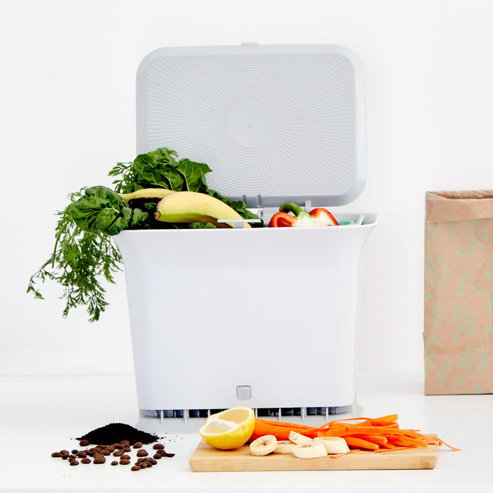A Full Circle Fresh Air Compost Bin sits open on the counter, filled with kitchen scraps. In front, coffee grounds, banana slices, and carrot sticks rest on a cutting board with a paper bag close by.