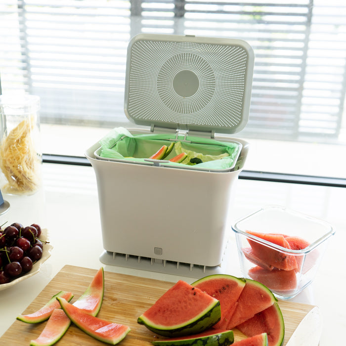 On a kitchen counter sits the Full Circle Fresh Air Compost Bin, filled with watermelon rinds and food scraps. Nearby are cherries, a container of cut watermelon, and a cutting board with sliced watermelon. Sunlight streams through window blinds.