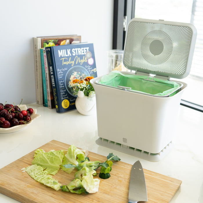 A Full Circle Fresh Air Compost Bin with its lid open sits on a kitchen counter near a window. Food scraps, cabbage leaves, and a knife are on the cutting board, while cookbooks and a bowl of cherries add color in the background.