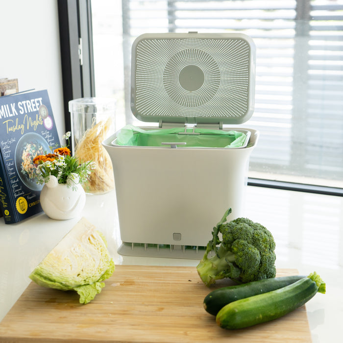 A Full Circle Fresh Air Compost Bin with its lid open sits on a kitchen counter by a cutting board of cabbage, broccoli, and zucchinis—an odor-free way to reduce kitchen waste. Nearby are a cookbook, jar, and vase of flowers by the window.