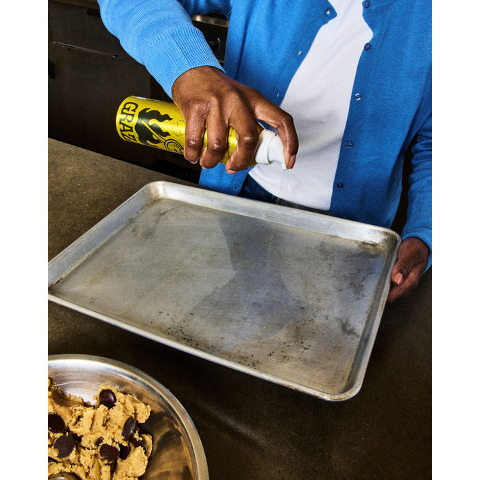 In a kitchen, someone wearing a blue sweater sprays Grazas Frizzle High Heat Cooking Oil from a squeeze bottle onto a metal baking tray. A bowl of chocolate chip cookie dough is partially visible on the countertop in the foreground.