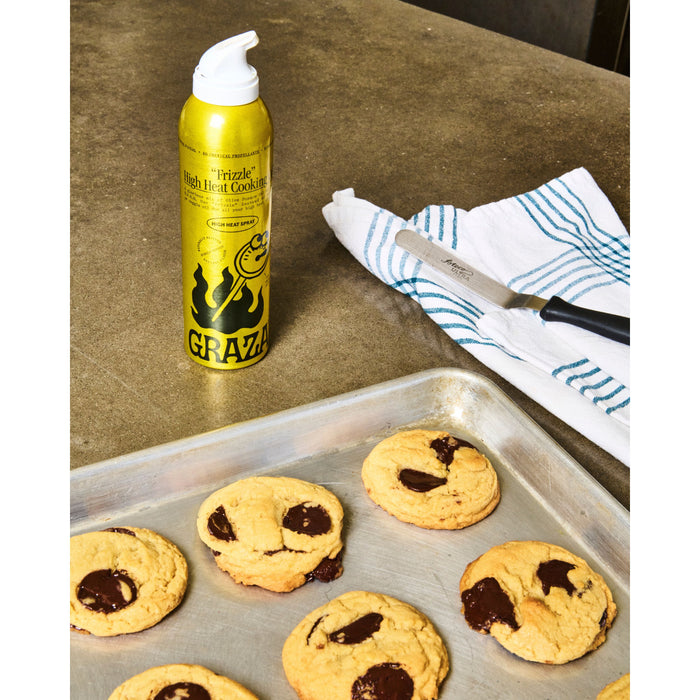A baking sheet with freshly baked chocolate chip cookies rests on a countertop. Beside it is a bottle of Grazas Frizzle High Heat Cooking Oil, ideal for high-temperature cooking, and a striped dish towel with a knife on top.