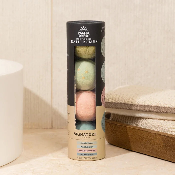 A cylindrical package of Pacha Soap Co. Froth Bomb, Pacha Peaces bath bombs, featuring four pastel-colored bombs, sits on a bathroom counter beside folded towels.