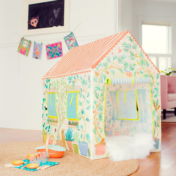 The DJECO Garden Tent Play House, resembling a floral-patterned cottage, sits on a woven rug and is ideal for imaginative play. Nearby, a play tea set and basket complete the kids hideout. Art displayed on a string against the white wall brightens the room.