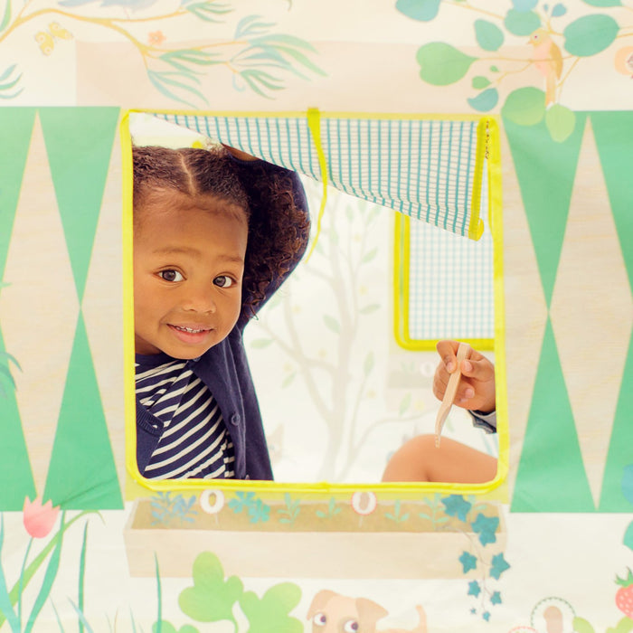 A child with curly hair and a striped shirt smiles while peering through the Garden Tent Play House by DJECO. Its playful design, featuring green patterns and floral elements, makes it perfect for a kids hideout. Another hand is visible, holding a toy thermometer.