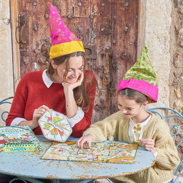 A woman and girl in party hats enjoy the Gathering a Garden Board Game by eeBoo Piece & Love, smiling as they play at a metal table. The vibrant game box sits beside them while they move pieces on the board.