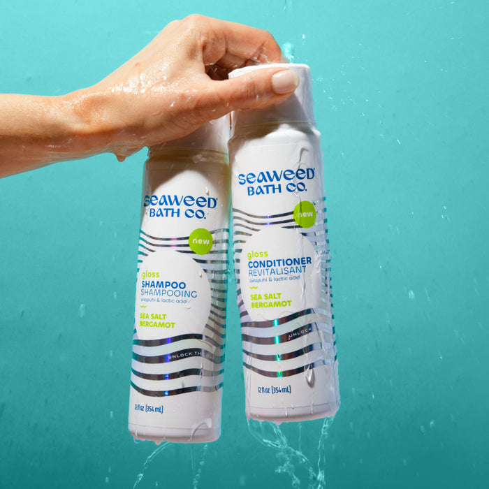 A hand holds two bottles from The Seaweed Bath Co under running water: Gloss Shampoo and Gloss Conditioner, featuring sea salt and bergamot with a replenishing moisture blend against a light blue backdrop with blue and green designs.