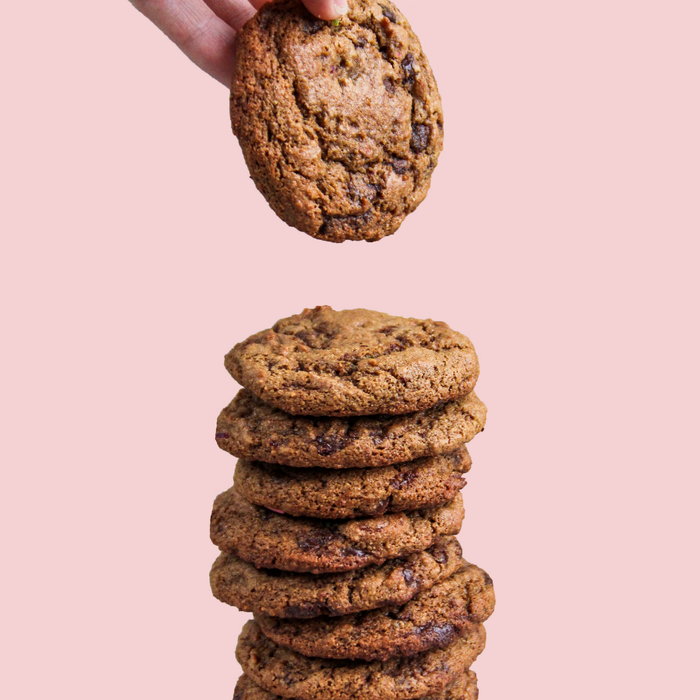 A hand adds a cookie to a stack of almond flour-made Stellar Eats Grain-Free Chocolate Chip Cookies, set against a light pink background for a playful touch.