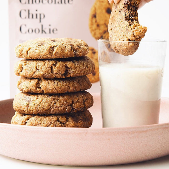 A pink plate holds a stack of cookies made from Stellar Eats Grain-Free Chocolate Chip Cookie Mix. Nearby, a hand dips another cookie into milk. In the background is the box, highlighting its almond flour base.