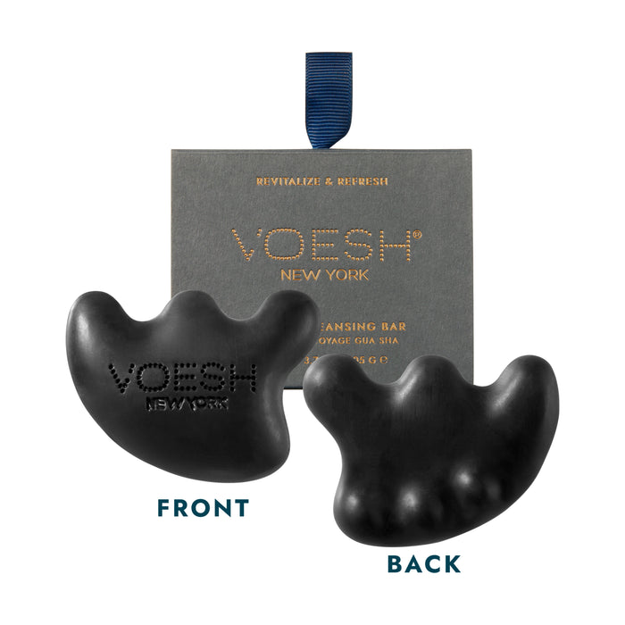 Image of a VOESH New York cleansing bar in black, shaped uniquely with four rounded protrusions. The front and back of the bar are displayed. The front view shows "VOESH NEW YORK" embossed on it. Behind the bars, there's packaging with a blue ribbon.