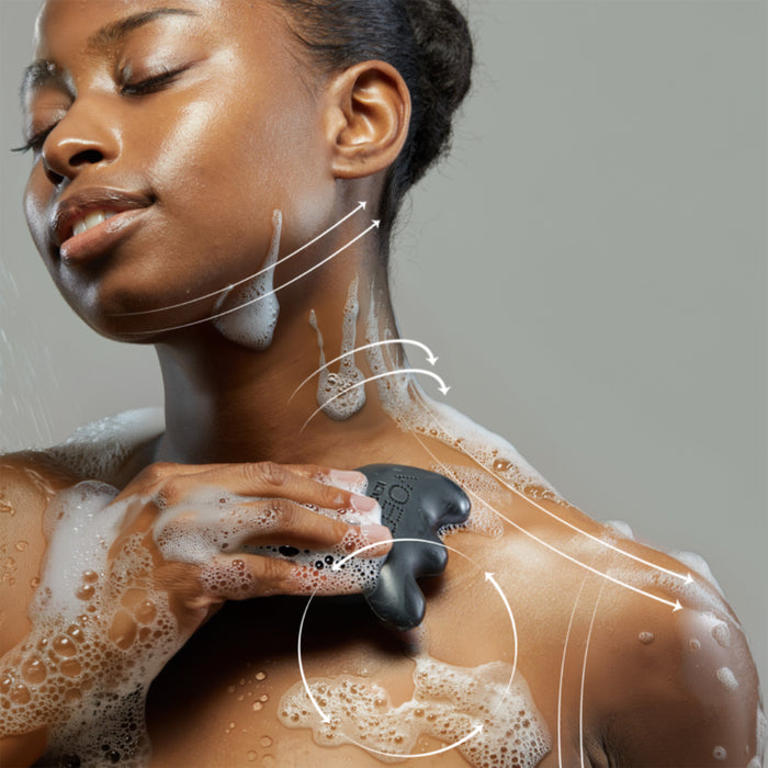 A woman uses a black exfoliating tool on her shoulder, covered in suds from VOESH New Yorks Gua Sha Cleansing Bar. White arrows indicate motion as the tool glides effortlessly, revealing detoxified skin. She looks relaxed while showcasing her skincare routine against a neutral background.