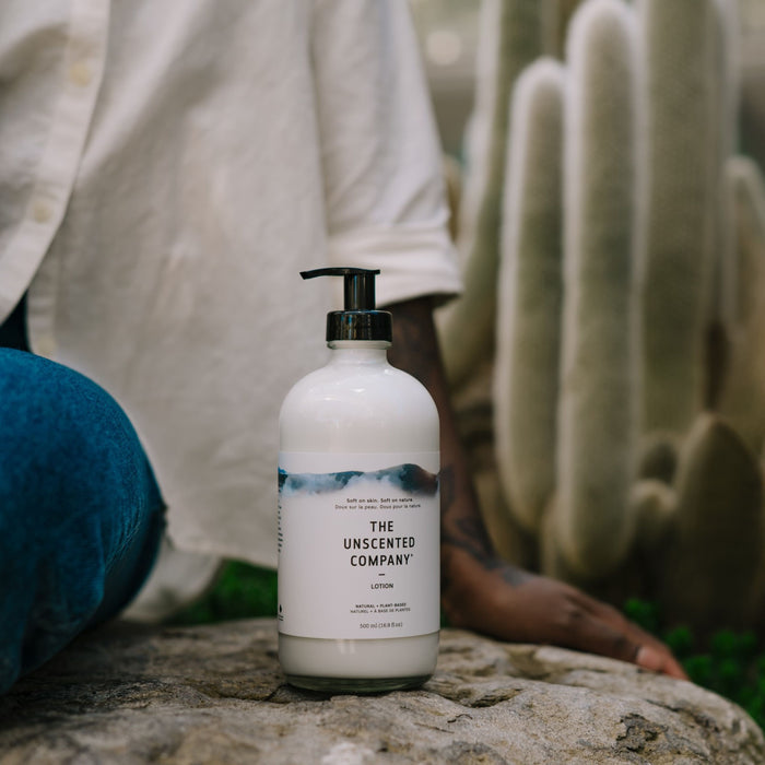 A person in a white shirt and blue jeans sits by a rock holding The Unscented Company Hand and Body Lotion, crafted from naturally derived ingredients. A large cactus adorns the background, enhancing the serene, earthy ambiance.