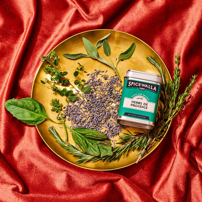 A gold plate on red fabric displays fresh ingredients, dried lavender, and a Spicewalla Herbs de Provence tin. The herb blend and tin are arranged to create an appealing, vibrant showcase.