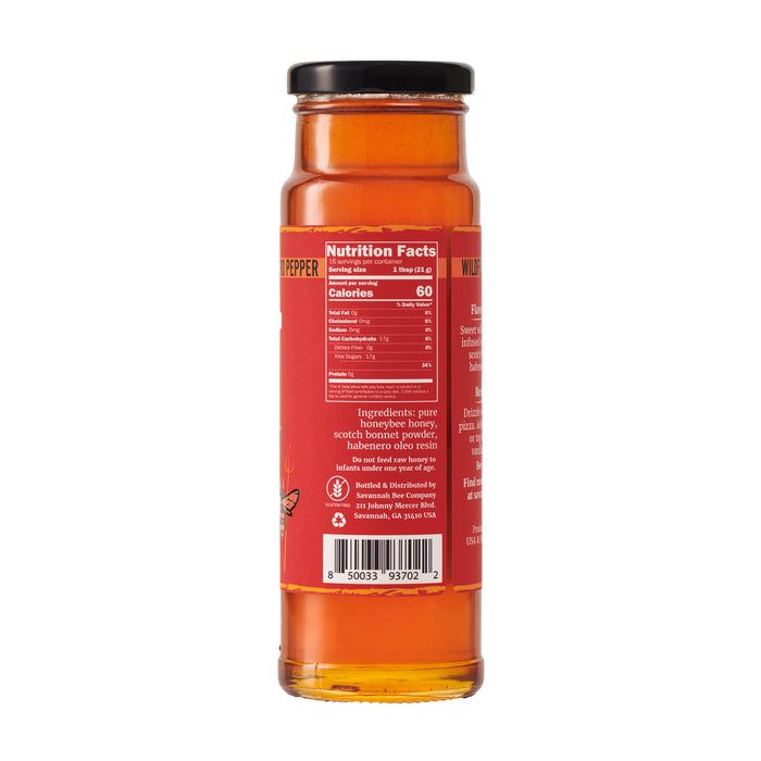 A tall glass jar with a black lid contains fiery Hot Honey sauce from Savannah Bee Company. The red label, featuring white text, lists nutrition facts and ingredients such as acacia honey, scotch bonnet pepper, and habanero peppers. A barcode is near the bottom.