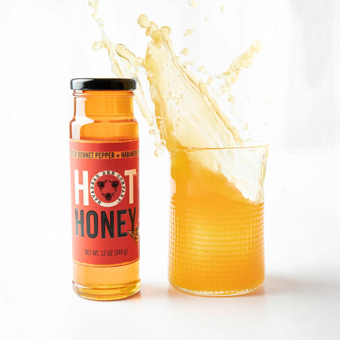 A bottle of Savannah Bee Company Hot Honey, made with acacia honey, scotch bonnet, and habanero peppers, is beside a glass filled with the same vibrant honey. Liquid splashes out against a clean white background.