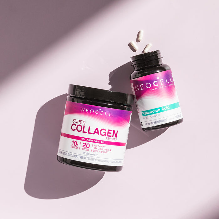 Two NeoCell bottles—Super Collagen and Hyaluronic Acid, 100 mg—sit on a pink surface with three white capsules nearby. Bright lighting creates soft shadows, highlighting the promise of a hydrated complexion.