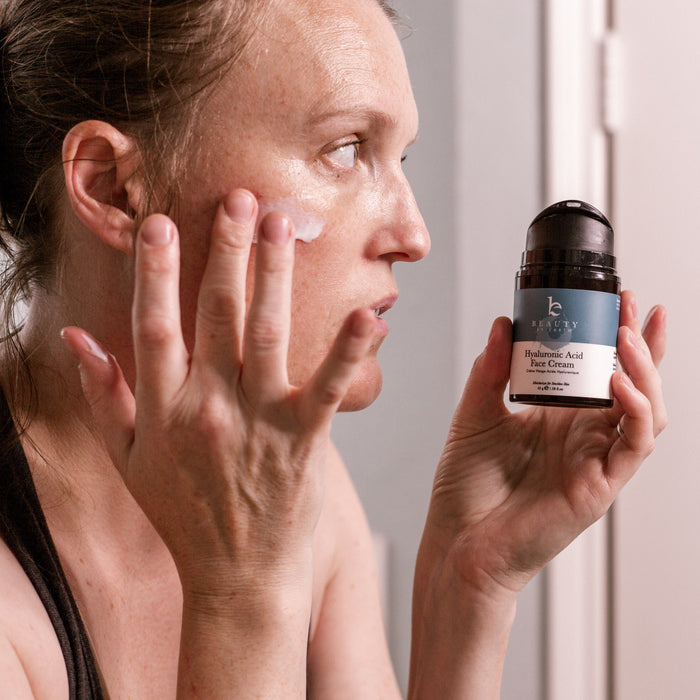 A woman, wearing a black tank top, gazes into the mirror as she applies a small amount of Beauty by Earths Hyaluronic Acid Cream to her cheek with her fingers, holding the bottle in her other hand.