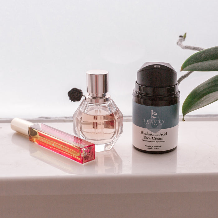 A perfume bottle, a long rectangular lipstick, and Beauty by Earths black cylindrical Hyaluronic Acid Cream sit on a white countertop. A green leaf is partially visible on the right side.