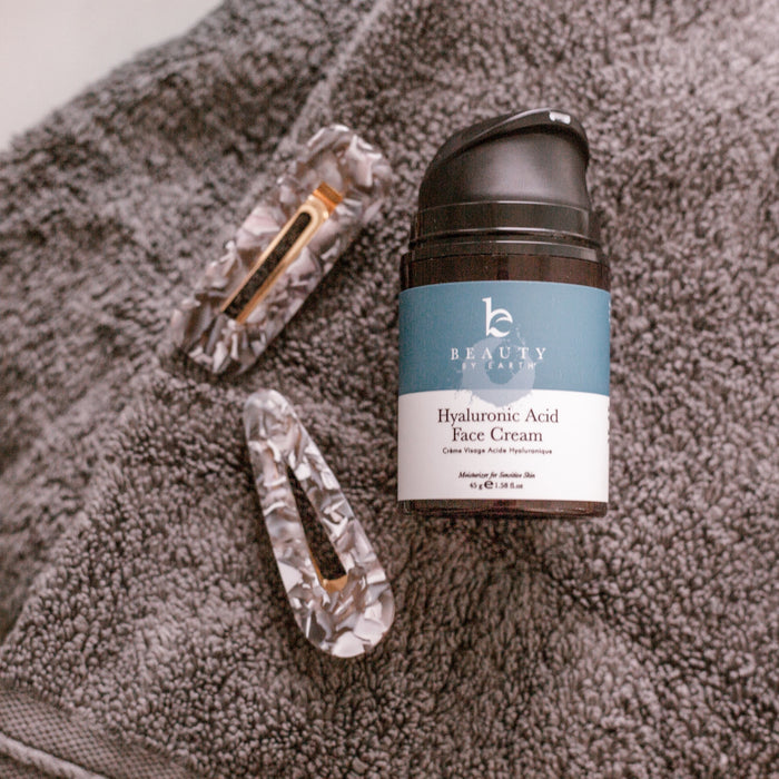 A bottle of Beauty by Earths Hyaluronic Acid Cream sits on a gray textured towel. Beside it are two marbled hair clips with gold accents, creating an inviting scene that promises a rejuvenating skincare and beauty routine.