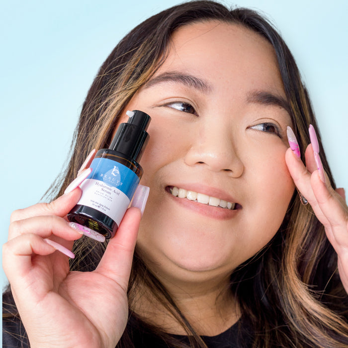 A person with long hair and pale pink nails smiles, holding a small bottle of Beauty by Earth’s Hyaluronic Acid Serum near their face. The serum features a black pump and blue label promising a youthful glow, set against a light blue background.