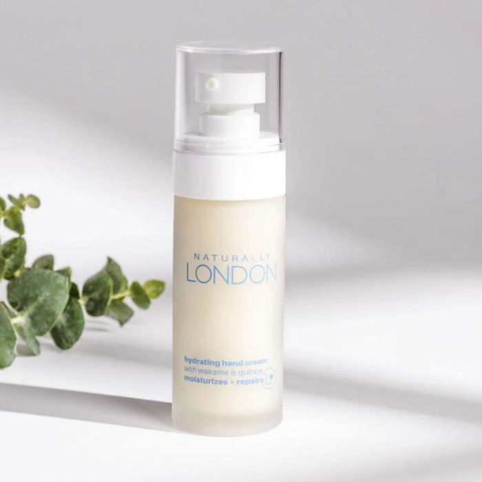 The Naturally London Hydrating Hand Cream, in a white bottle with a clear pump top, rests against a light backdrop. Infused with wakame bio-ferment and quinoa protein for nourishment and hydration, its artfully paired with green leaves to highlight its natural allure.