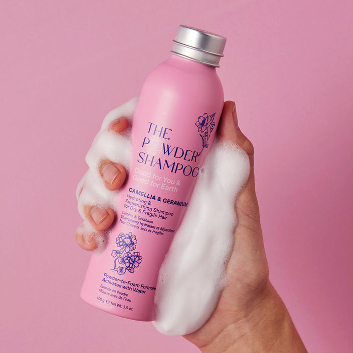 A hand holds a pink cylindrical bottle of The Powder Shampoos Hydrating & Replenishing Powder Shampoo against a pink background. Rich foam showcases its powder-to-foam formula with nourishing Camellia and Geranium, ideal for dry or chemically treated hair.