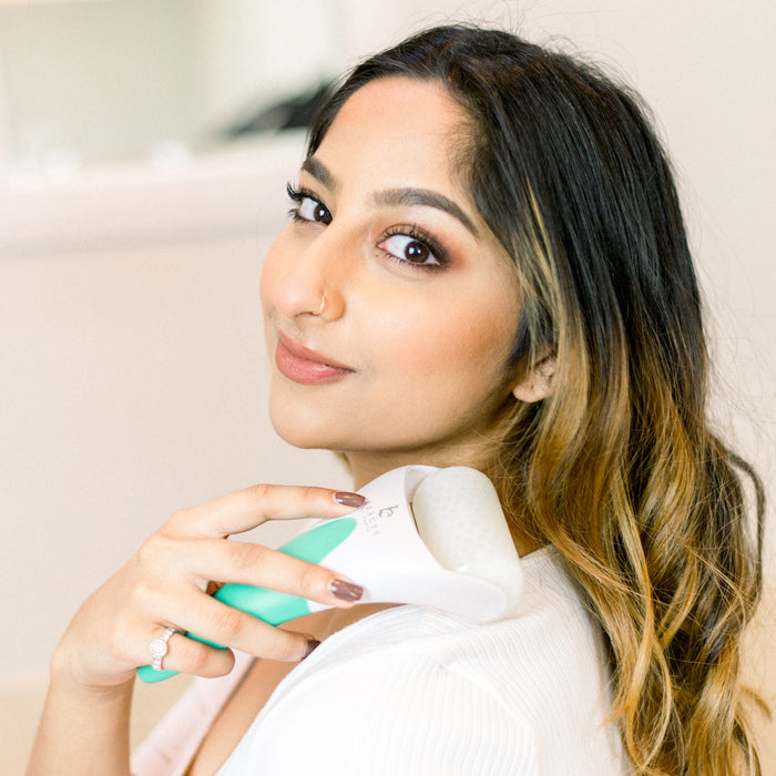 A long-haired person with a nose ring uses Beauty by Earths Ice Roller on their neck for lymphatic drainage. They wear a white top, and the roller’s green handle and white head enhance its natural beauty appeal against a softly blurred background.