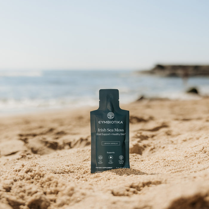 A packet of Cymbiotika Irish Sea Moss stands upright on sandy beach with ocean and blue sky blurred in the background, emphasizing its gut health benefits.